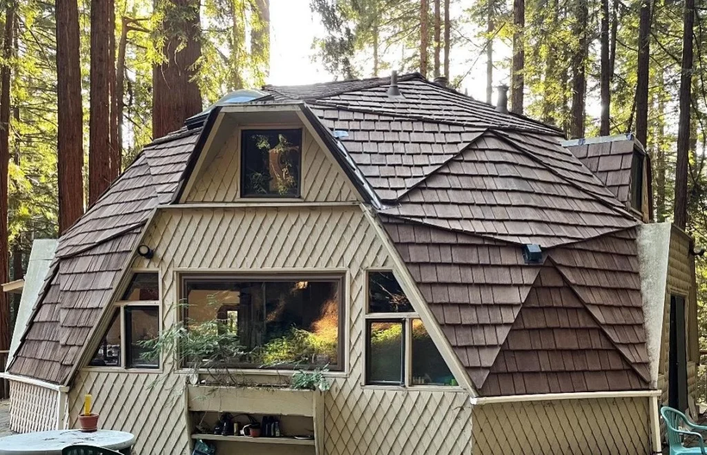 Geometric cabin with cedar shake roofing nestled among tall forest trees.