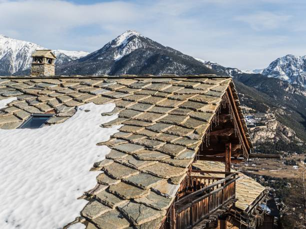 Stone tile roof with patches of snow on a mountain cabin surrounded by alpine scenery.