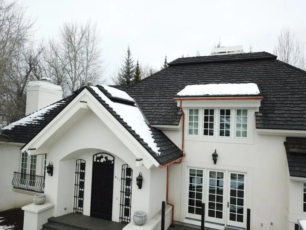 Dark composite shake roof with snow coverage and copper gutters on a residential home.